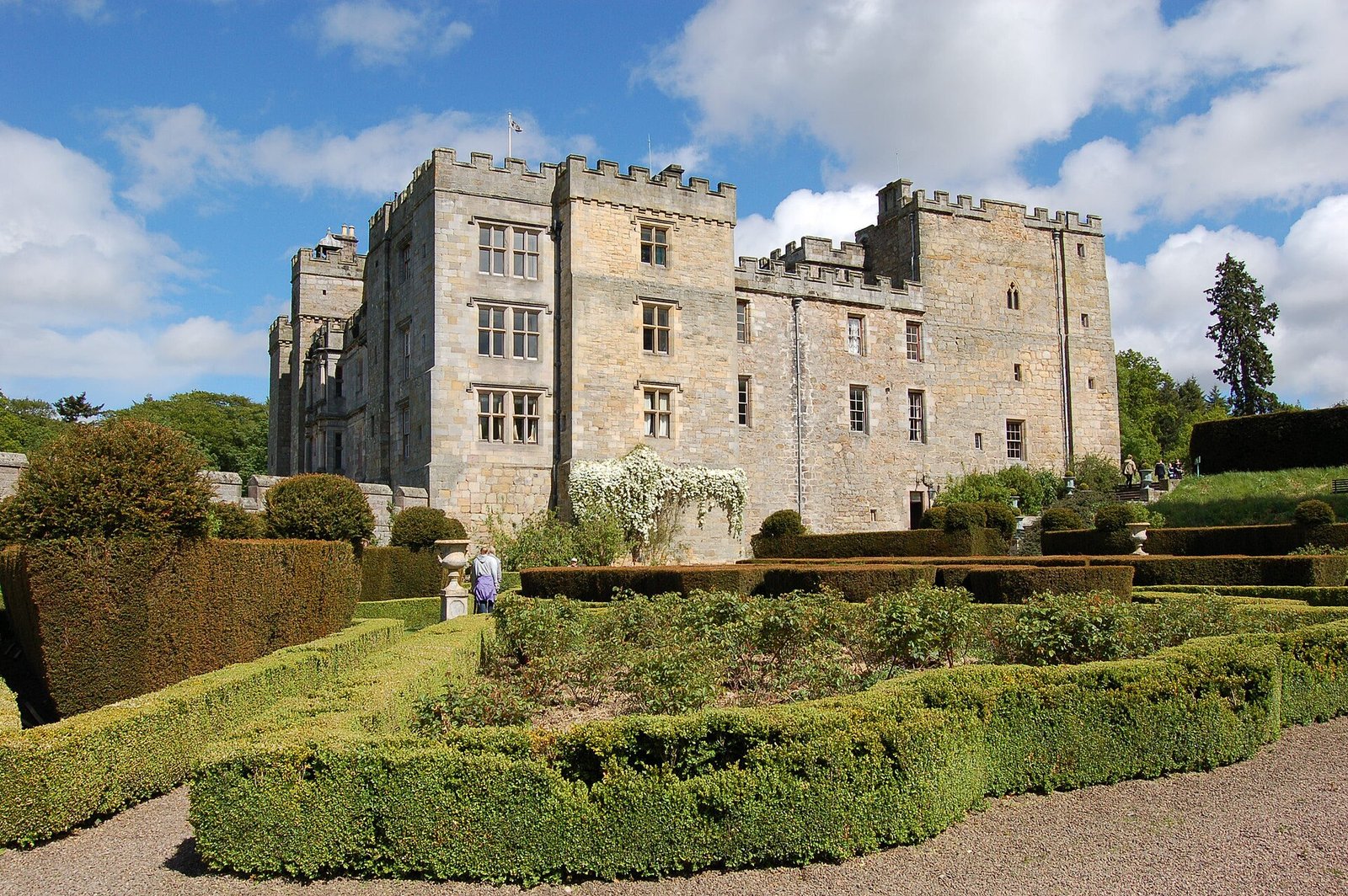 Vista exterior del castillo de Chillingham, con torres de piedra almenadas y jardines geométricos en primer plano.