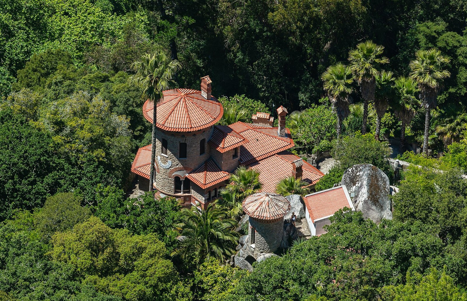 Vista elevada de la Vila Sassetti, con tejados de teja roja, una torre circular y grandes rocas entre la vegetación de Sintra.