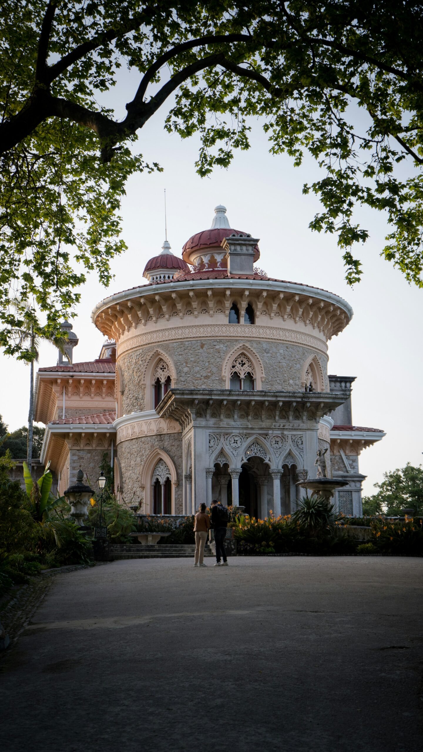 Fachada circular de piedra del Palacio de Monserrate, con arcos ornamentados, cúpulas rojizas y dos visitantes bajo los árboles.