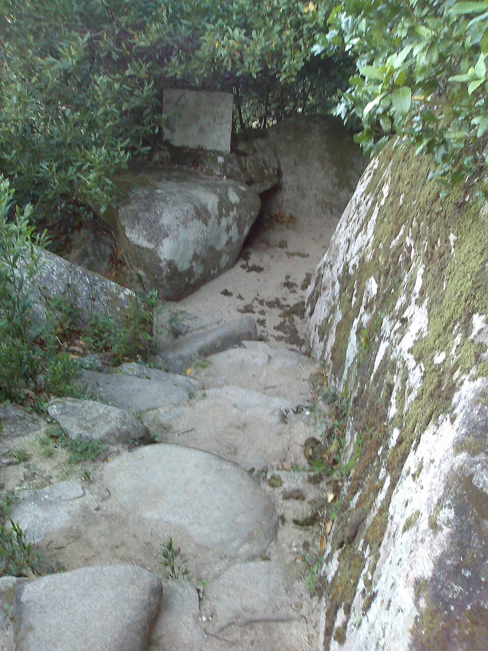 Escalones de piedra que descienden entre grandes rocas de granito y vegetación hacia la Cova de Frei Honório, en Sintra.